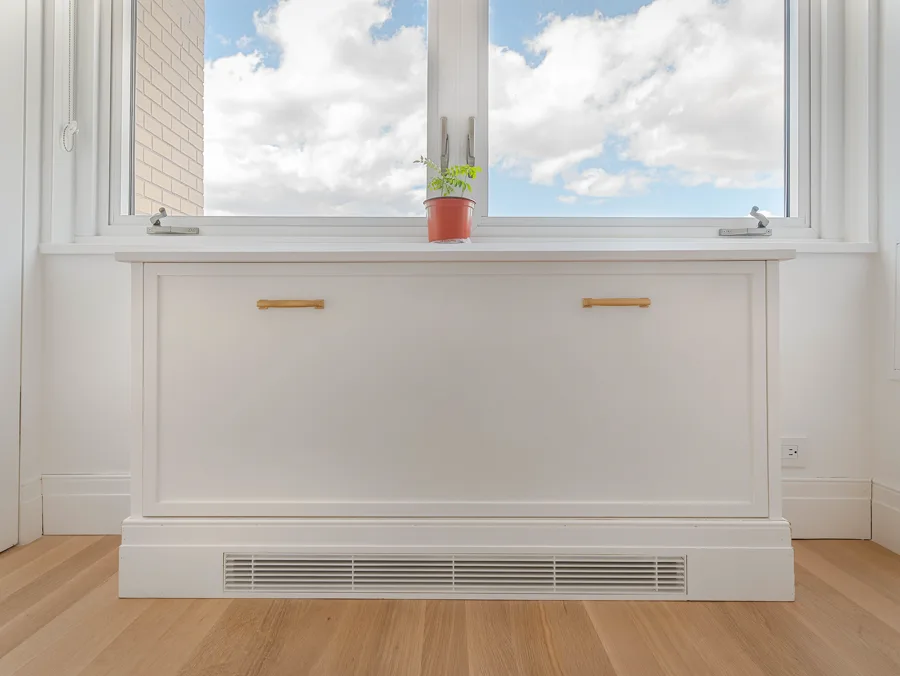 White built-in cabinet with brass pulls fitted under a bay window on an oak floor
