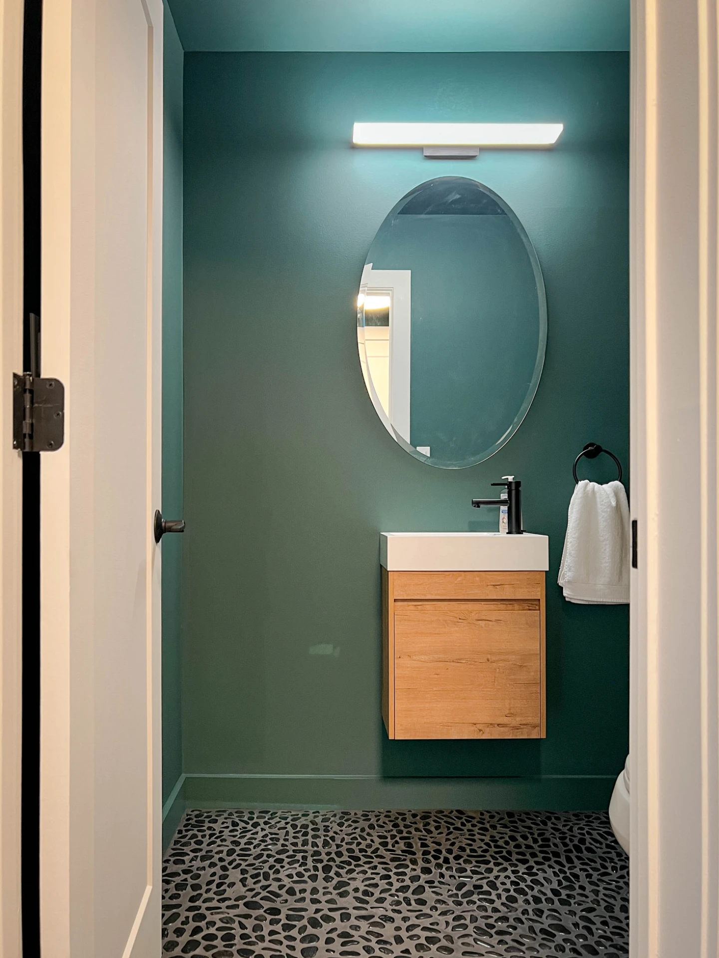 Small powder room painted teal, with an oval mirror and a matte black faucet